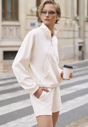 Cremefarbener Zip-Up-Sweatshirt und passende Shorts, aus weichem Stoff mit lässigem Schnitt. Das Model hält eine Kaffeetasse und trägt eine Sonnenbrille.