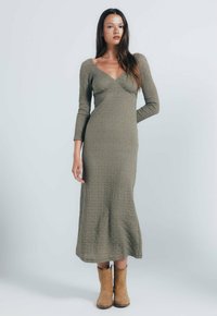 Langes olivengrünes Kleid aus strukturiertem Stoff, mit V-Ausschnitt und langen Ärmeln, kombiniert mit beigen Cowboy-Stiefeln. Bodengängiges Design.