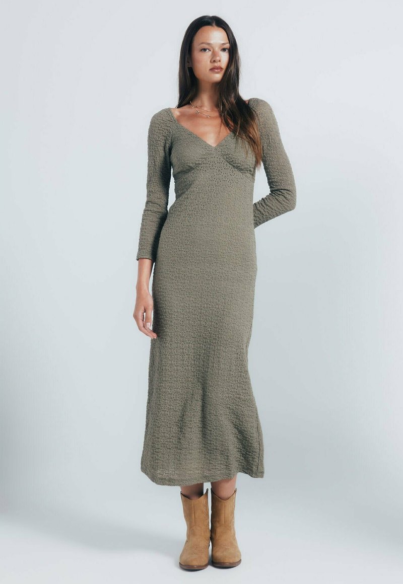 Langes olivengrünes Kleid aus strukturiertem Stoff, mit V-Ausschnitt und langen Ärmeln, kombiniert mit beigen Cowboy-Stiefeln. Bodengängiges Design.
