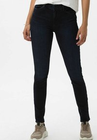 Mörkblå skinny jeans med standard midja, fem fickor och lätt stretch; bärs med beige sneakers och en vit topp. Slät textur.