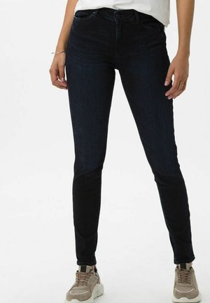 Jean skinny bleu foncé avec taille standard, cinq poches et léger stretch ; porté avec des baskets beiges et un haut blanc. Texture lisse.