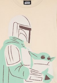 T-shirt beige clair avec un graphique d'une figure casquée en vert menthe, tenant un petit personnage vert, avec des contours foncés et un design simplifié.