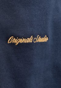 Υφάσμα σε ναυτικό μπλε με μπεζ κειμενο που είναι κεντημένο με καλλιγραφικά γράμματα που γράφουν "Originals Studio" σε πλεκτή υφαντική επιφάνεια.