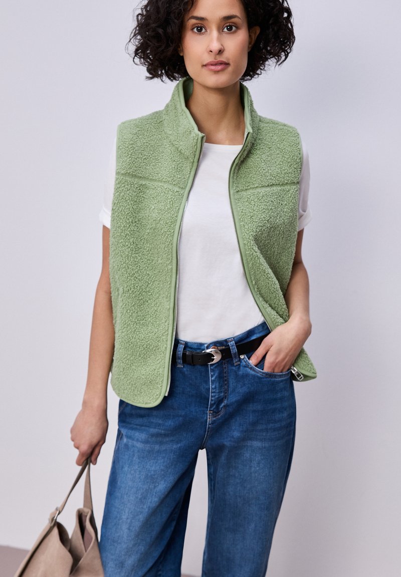 Frau trägt hellgrüne, strukturierte Weste über weißem T-Shirt und blauer Jeans, hält eine beige Tragetasche und steht vor einem einfarbigen, hellen Hintergrund.