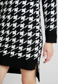 Pletené šaty s černobílým vzorem houndstooth, dlouhými rukávy a bočními rozparky na dolním lemu pro pohodlný pohyb.