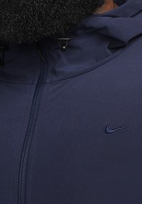Gros plan sur la fermeture éclair et la capuche d'une veste bleu foncé, avec un petit logo Nike brodé sur la poitrine et une partie d'une barbe au-dessus du col.