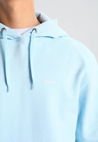 Sweat à capuche bleu clair en tissu doux, avec un logo "VANS" minimaliste brodé en rose, des cordons réglables et une capuche confortable.
