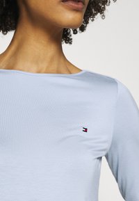 Primo piano di una persona che indossa una camicia a maniche lunghe di colore azzurro chiaro con un piccolo logo rettangolare rosso, bianco e nero sul petto.