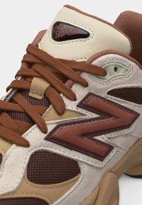 Κοντινό πλάνο του αθλητικού παπουτσιού New Balance 9060 με καφέ κορδόνια, μπεζ και λευκά πάνελ και μεγάλο καφέ λογότυπο "N" στο πλάι.