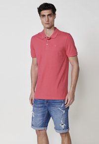 Roza polo majica s tremi gumbi in majhnim logotipom, v kombinaciji z modrimi denimski kratkimi hlačami z uničenimi detajli in zavitimi robovi.