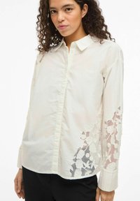 Balta krekls ar ziedu izšuvumiem caurspīdīgajās piedurknēs, ar apkakli, pogām priekšpusē un gludu, teksturētu audumu.