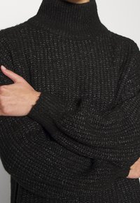 Svart stickad tröja med hög polo, med ett strukturerat mönster och vida ärmar. Mjuk, bekväm material med en avslappnad passform.