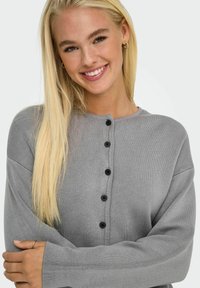 Grå stickad cardigan med rund hals, svarta knappar framtill och vida ärmar. Texturen är mjuk och något elastisk.