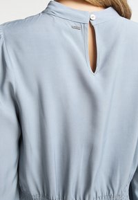 Rückansicht einer Person in einer hellblauen Bluse mit einem durch Knöpfe versehenen Ausschnitt und gerafftem Taillendetail.