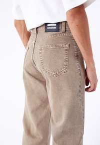 Vaqueros de mezclilla beige con un ajuste relajado, que cuentan con bolsillos traseros y una etiqueta de marca negra en la cinturilla. Textura suave y costuras resistentes.