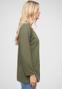 Khaki green long-sleeve blouse in a textured, ribbed fabric. Relaxed fit with a V-neck and straight hem. Paired with blue jeans.