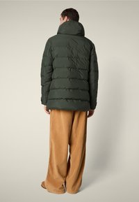 Uomo di spalle che indossa un giubbotto imbottito verde scuro con colletto alto e pantaloni in corduroy beige a gamba larga.