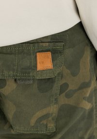 Cargo kalhoty v zeleném maskovacím vzoru, s koženou nášivkou "Alpha Industries", kontrastním šitím a kapsou s klopou.