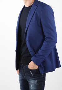 Blazer blu con texture e rever a punta. Indossato sopra un maglione nero, abbinato a jeans scuri strappati. Giacca aperta che mostra la tasca interna.