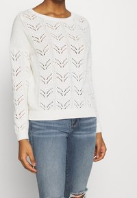 White knitted sweater with a chevron pattern, long sleeves, and ribbed cuffs, paired with light blue distressed denim shorts.