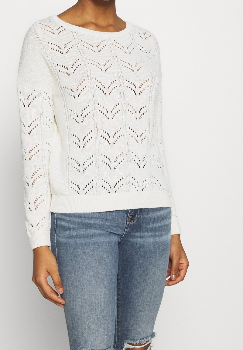 White knitted sweater with a chevron pattern, long sleeves, and ribbed cuffs, paired with light blue distressed denim shorts.