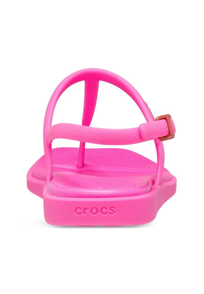 Różowe gumowe sandały z płaską podeszwą, z dwoma wąskimi paskami i małym metalowym zapięciem. Nazwa marki "Crocs" widoczna z boku.