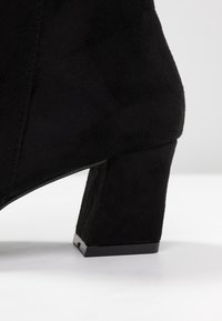 Sorte ankelstøvler lavet af ruskind med en blokhæl, der har en glat overflade, rene linjer og en skinnende sort sål.