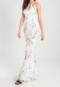 Abito maxi bianco a fiori con scollatura a occhiello, corpino fitted, gonna fluida e dettagli in pizzo sul fondo. Si indossano sandali neri.