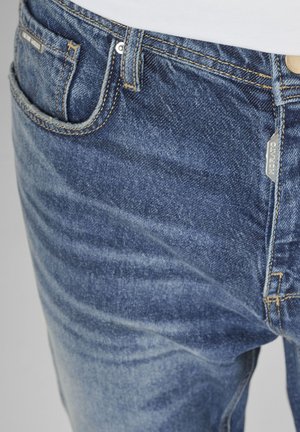 Detaljni prikaz plavih traperica koji pokazuje prednji džep, omče za remen, metalne zakovice i oznaku s logotipom na pojasu i šavu džepa.