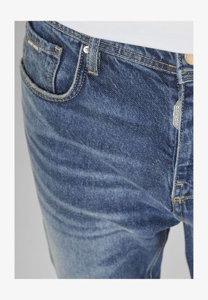 Detaljni prikaz plavih traperica koji pokazuje prednji džep, omče za remen, metalne zakovice i oznaku s logotipom na pojasu i šavu džepa.