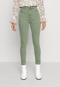 Jeans verdes de tiro alto y corte ajustado con una textura suave y un clásico diseño de cinco bolsillos, combinados con botas de tobillo blancas y brillantes.