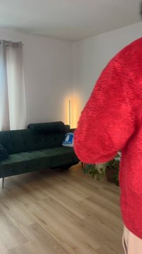 Άτομο που φοράει κόκκινο πουλόβερ με υφή που κρατά ένα ποτήρι, στέκεται κοντά σε ένα σκούρο πράσινο καναπέ με μαξιλάρια σε ένα φωτεινό σαλόνι.