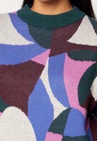 Neulepusero, jossa on vihreä ribbireunus ja monivärisiä geometrisia kuvioita purppurana, vaaleanpunaisena, valkoisena ja tumman vihreänä.