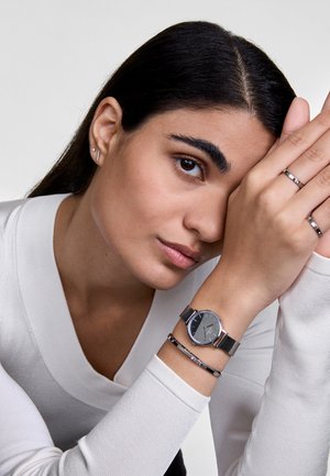 Montre en mailles argentées avec un cadran gris foncé, accompagnée de fines bagues polies et d'un bracelet avec des détails subtils, sur un fond de pull clair.