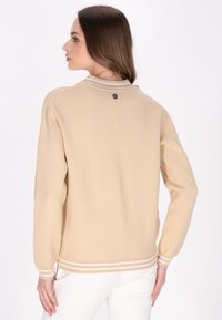 Beige stickad tröja med rundad halsringning, vita randiga detaljer vid ärmslut och nederkant, samt en metallisk logodetalj på baksidan.