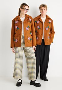 Καφέ σακάκια από μάλλινο μείγμα με κεντητά ροζ λουλούδια και πράσινα φύλλα. Συνδυασμένα καρό παντελόνια με φαρδιά πόδια, μαύρα παπούτσια και γυαλιά.