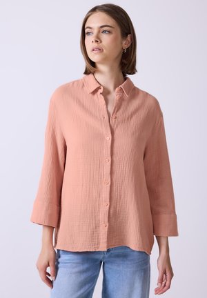 Camicia color pesca con bottoni, realizzata in tessuto testurizzato e sgualcito, colletto e maniche a tre quarti. Indossata con jeans azzurri chiari.