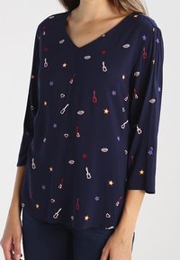 Un haut bleu marine à col en V avec des manches 3/4, comportant des motifs brodés de guitares, d'étoiles et de lèvres dans diverses couleurs sur un tissu doux.