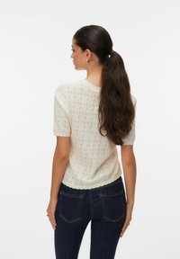 Kreemika värvi lühikeste varrukatega särk tekstuurse mustri, lainelise dekolteega ja keha järgiva lõikega, kombineeritud tumeda teksapükstega.