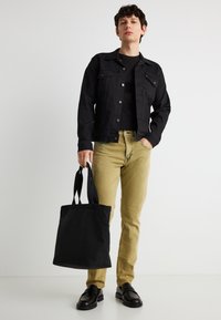 Νεαρός άνδρας με μαύρο μπουφάν και καφέ τζιν, κρατώντας μαύρη τσάντα ώμου στο δεξί χέρι, στέκεται μπροστά σε ένα λευκό φόντο.