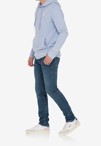 Sudadera de forro polar azul claro con un bolsillo frontal y logo, combinada con jeans oscuros de corte slim y zapatillas blancas con acentos azules.