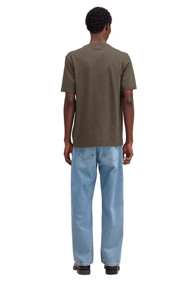 Mężczyzna z krótkimi, kręconymi włosami, ubrany w oliwkowo-brązowy t-shirt z krótkim rękawem, jasnoniebieskie dżinsy i czarne buty, stoi tyłem.