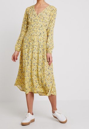 Femme portant une robe midi jaune à motifs floraux avec manches longues et col en V, associée à des baskets à plateforme blanches, debout devant un fond uni.