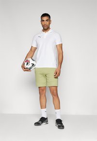 Camisa polo branca, calções verdes claros, sapatilhas de desporto pretas e meias brancas. Segurando uma bola de futebol estampada. Design simples e atlético.