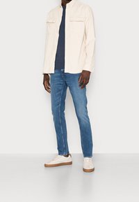 Camisa beige claro abotonada con dos bolsillos delanteros, camiseta azul marino debajo, vaqueros azules y zapatillas blancas con detalles en marrón.