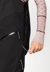 Melna kombinezona ar rāvējslēdzēja kabatām, piedāvājot matētu un spīdīgu audumu tekstūru. Modeļa roka ar rakstainu garo piedurkņu apģērbu pielāgo rāvējslēdzēju.