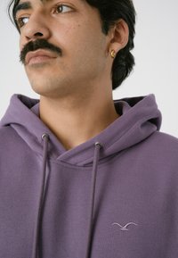 Lila Baumwoll-Hoodie mit Kapuze und einem kleinen gestickten Logo auf der Brust. Glatte Textur mit lockerer Passform.