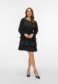 Juoda suknelė su sluoksniuotais raukiniais, ilgomis pūstomis rankovėmis ir aptemptu liemeniu. Derinama su mėlynais aukštakulniais su siaurais priekiu. Lygus audinys.