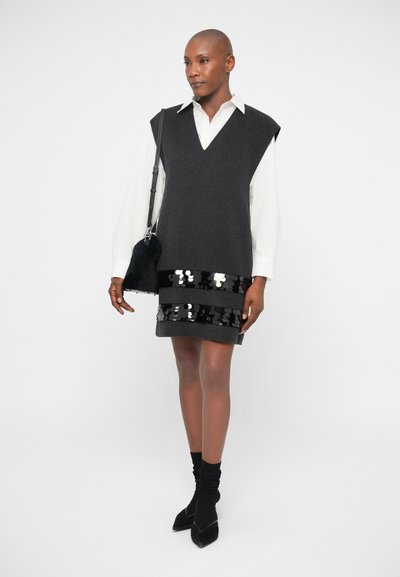 Vestido sem mangas cinza escuro com decote em V e detalhes em lantejoulas pretas. Estilizado com uma camisa branca de colarinho e acessórios pretos, incluindo uma bolsa pequena.