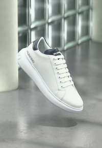 Λευκό δερμάτινο αθλητικό παπούτσι Valentino με μαύρη ταμπέλα στη φτέρνα και λογότυπο στο πλάι και στη γλώσσα, αιωρούμενο πάνω από ανακλαστικό γκρι δάπεδο και τοίχο από γυάλινα τούβλα.